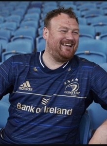 production manager Eoin Kilkenny seated in blue football stadium seats, wearing a navy leinster rugby jersey