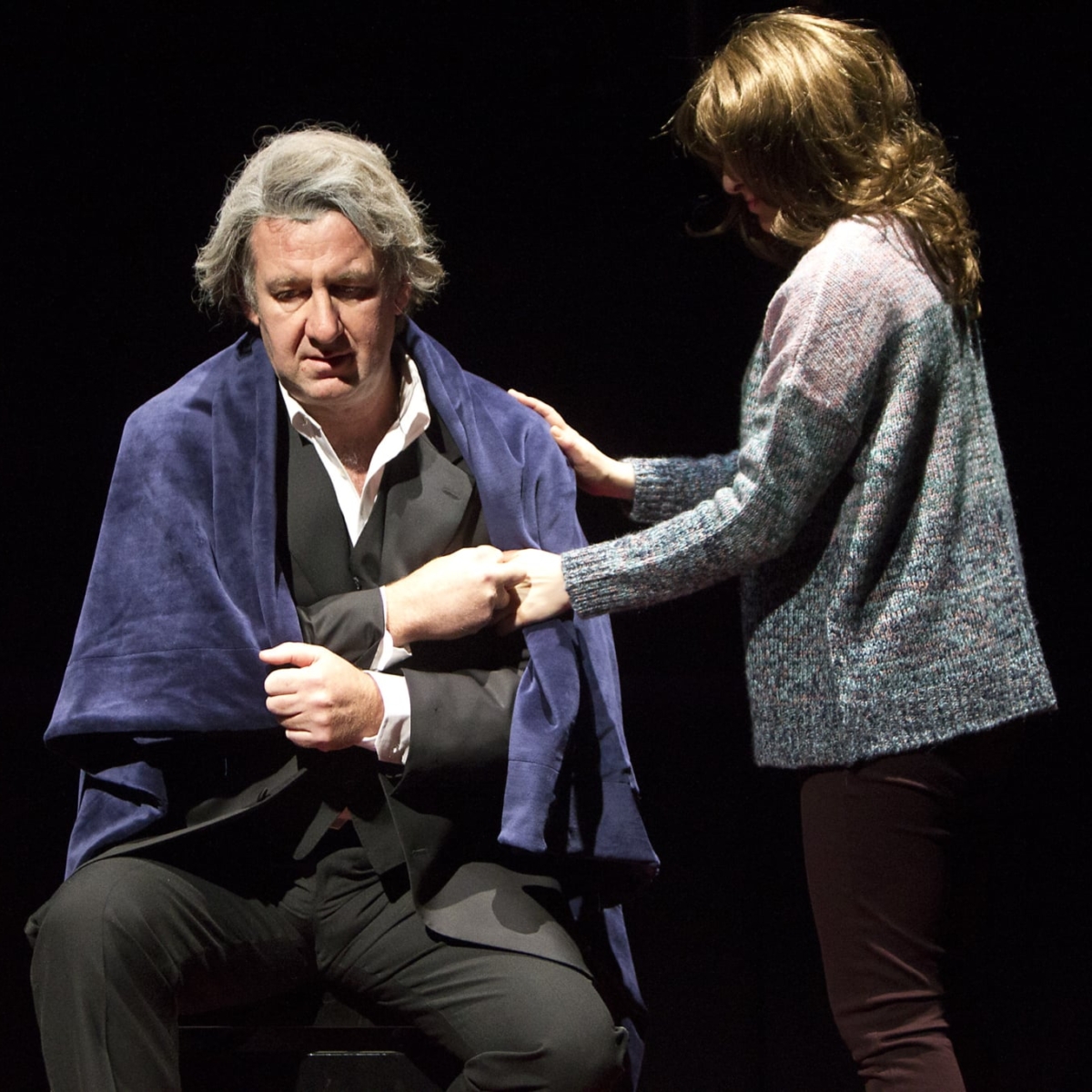 A young girl comforts an older man who has a blanket over his shoulders.