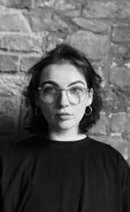 Black and white image of Emily Waters. Emily has dark hair, rounded glasses and is wearing hoop earrings.