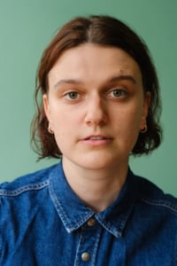 Catriona Faint headshot. Catriona has short brown hair that comes to just above their shoulders and is wearing a blue denim shirt. The background is green.