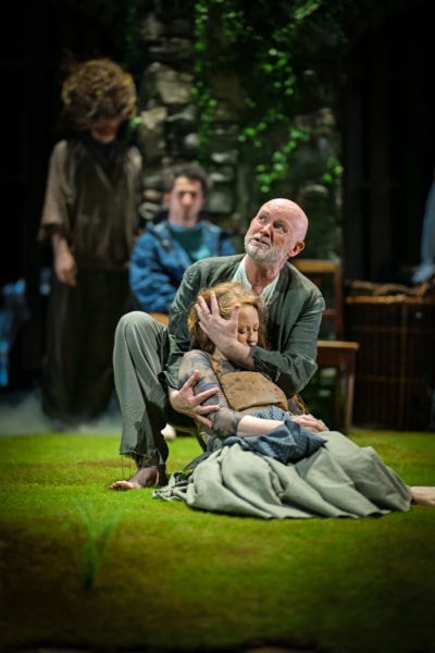 REFUGE production image. Herr Ireland played by Aidan Kelly kneels on the green grass floor holding actress Hannah Von Peinen who plays Hannah in his arms.