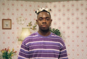 Artist Dafe Orugbo's headshot. He is wearing a purple t-shirt and standing in front of a pink wall with floral wallpaper.