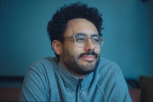 Seán Gallen's headshot. Sean is wearing a grey/blue zip up fleece and transparent rimmed glasses. He is photographed in from of a blue backdrop.