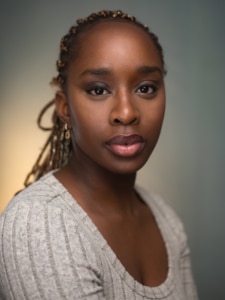 Tishé Fatunbi Headshot. Tishé is wearing a cream top and is standing against a grey blue background that has a light source peaking through over her right shoulder.
