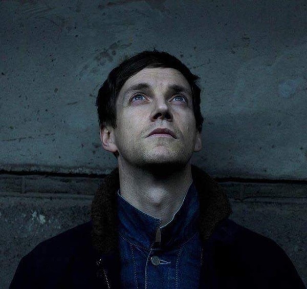 Writer and performer Emmet Kirwan, fstanding against a greay/blue concrete wall wearing a blue jumper and facing the sky
