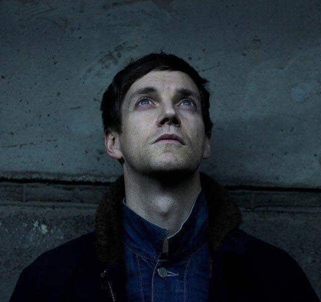 Writer and performer Emmet Kirwan, fstanding against a greay/blue concrete wall wearing a blue jumper and facing the sky