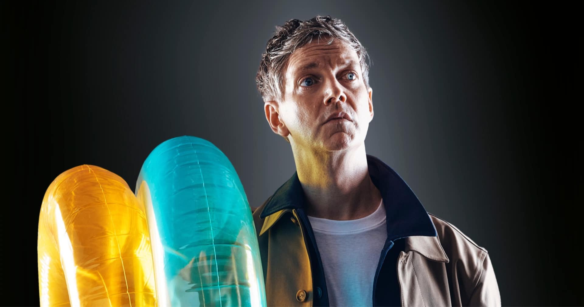 Emmet Kirwan, photographed from the chest up, wearing a beige jacket and a white tshirt and holding a yellow and turquoise floaty ring.